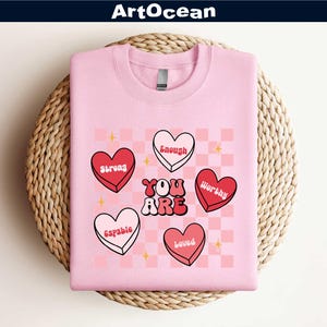 May include: Pink sweatshirt with the words "YOU ARE" in a retro font, surrounded by heart-shaped candies with words like "Strong" and "Worthy". The design is set against a pink and white checkered background with gold stars.