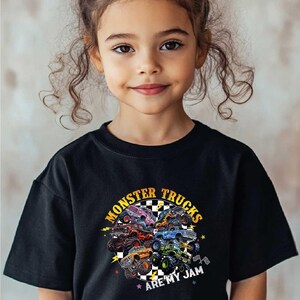 A young girl with curly hair wearing a black t-shirt with a graphic design of monster trucks and the text "Monsters Trucks Are My Jam" printed on it.