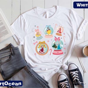 A white t-shirt with a colorful graphic design featuring various cartoon characters and snow globes, along with a pair of jeans, a book, a cup, and a pair of sneakers.