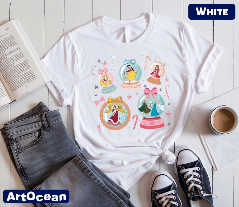 A white t-shirt with a colorful graphic design featuring various cartoon characters and snow globes, along with a pair of jeans, a book, a cup, and a pair of sneakers.