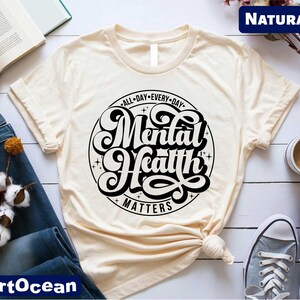 May include: Cream-colored t-shirt with the text "Mental Health Matters" in a black, circular design. The shirt is knotted at the waist and is paired with blue jeans and gray sneakers. A cup of coffee and an open book are also visible.