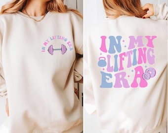 May include: A cream-colored sweatshirt with the text "In My Lifting Era" in pink and blue lettering. The front of the sweatshirt features a pink dumbbell graphic. The back of the sweatshirt features the same text with a pink kettlebell graphic.