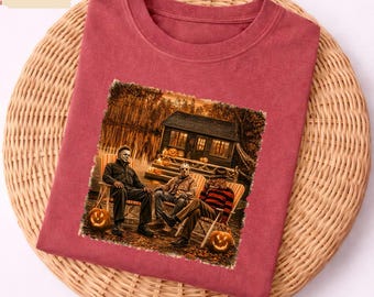 May include: A crimson crew neck t-shirt featuring a graphic print of three horror movie characters sitting on chairs in front of a cabin at night. The scene includes lit jack-o'-lanterns on the porch and ground, with a forest background.