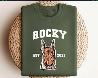 May include: An olive green sweatshirt with the name "ROCKY" in white block letters. Below the name is a graphic of a Doberman Pinscher dog with the text "EST. 2021". The sweatshirt is displayed on a woven, circular surface.