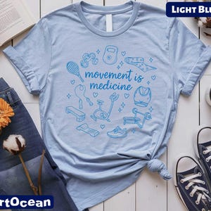 a light blue t-shirt with a graphic design of various fitness-related items, such as a tennis racket, dumbbells, and a heart, along with the text "motion is medicine". The t-shirt is displayed on a white background, accompanied by a pair of jeans, a pair of sneakers, a book, and a cup.