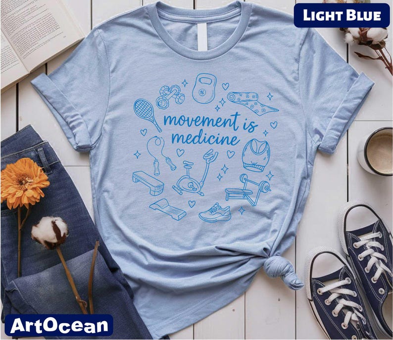 a light blue t-shirt with a graphic design of various fitness-related items, such as a tennis racket, dumbbells, and a heart, along with the text "motion is medicine". The t-shirt is displayed on a white background, accompanied by a pair of jeans, a pair of sneakers, a book, and a cup.