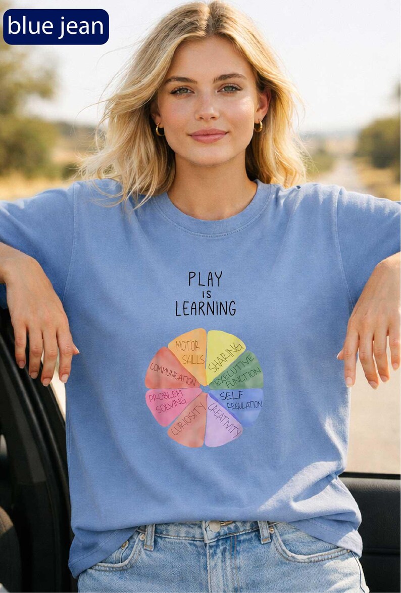 A woman wearing a blue t-shirt with a colorful wheel graphic and the text "PLAY IS LEARNING" printed on it.