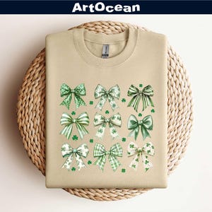 May include: A beige sweatshirt with a design of nine green and white bows with shamrock accents. The bows are arranged in three rows of three. The sweatshirt is on a woven wicker surface.