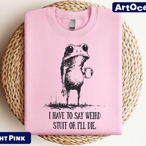 A pink t-shirt with a black and white illustration of a frog holding a cup, accompanied by the text "I HAVE TO SAY WEIRD STUFF OR I'll die" printed on it.