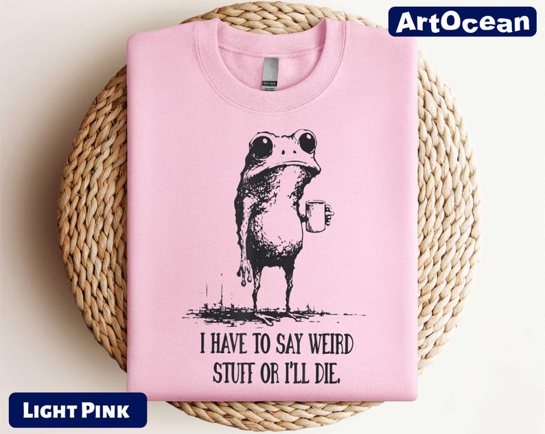 A pink t-shirt with a black and white illustration of a frog holding a cup, accompanied by the text "I HAVE TO SAY WEIRD STUFF OR I'll die" printed on it.