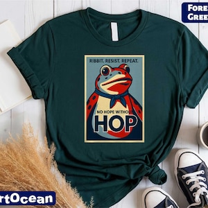 May include: Forest green t-shirt featuring a frog graphic with the text "Ribbit. Resist. Repeat." and "No Hope Without Hop." The frog is red, blue, and white. The shirt is paired with white sneakers and a coffee mug.