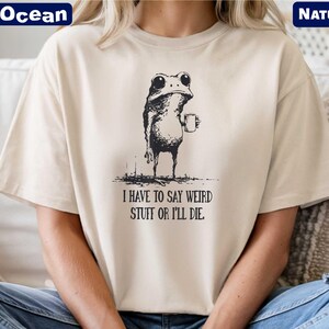 A person wearing a white t-shirt with a black and white illustration of a frog holding a cup and the text "I HAVE TO SAY WEIRD STUFF OR I'll die" printed on it.