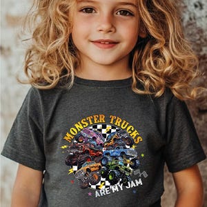 A young girl with curly blonde hair wearing a gray t-shirt with a graphic design of monster trucks and the text "MonSTER TRUCKS ARE MY JAM".