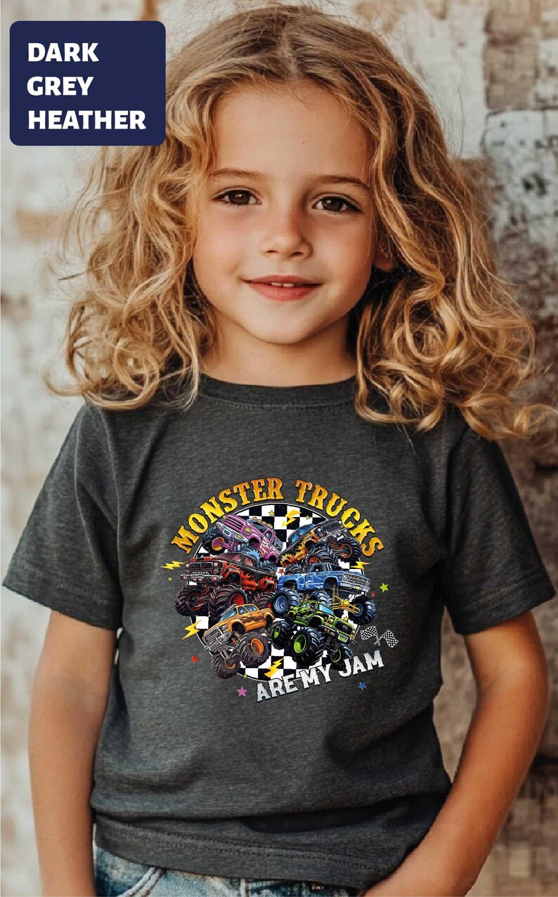 A young girl with curly blonde hair wearing a gray t-shirt with a graphic design of monster trucks and the text "MonSTER TRUCKS ARE MY JAM".