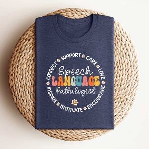 May include: A navy blue t-shirt with a white circle design. The circle features the text "Speech Language Pathologist" in rainbow colors with the words "Connect, Support, Care, Love, Inspire, Motivate, Encourage" surrounding the text.