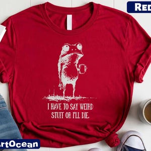 A red t-shirt with a white frog graphic and text, along with a pair of jeans and a cup of coffee, on a white surface.