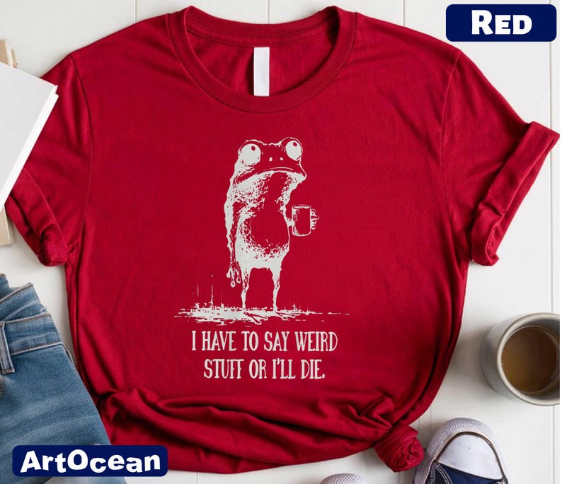 A red t-shirt with a white frog graphic and text, along with a pair of jeans and a cup of coffee, on a white surface.