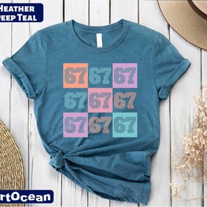 A blue t-shirt with a colorful graphic design of numbers on it, placed on a wooden surface alongside a hat and some dried flowers.