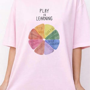 A woman wearing a pink t-shirt with a colorful wheel graphic and the text "PLAY IS LEARNING" printed on it.