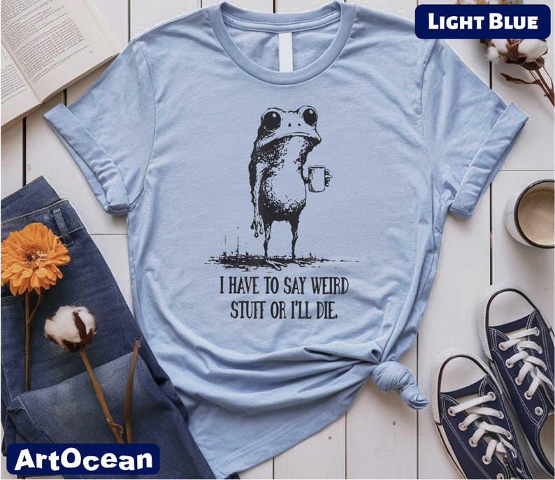 a light blue t-shirt with a graphic of a frog holding a cup, accompanied by the text "I have to say weird stuff or I'll die". The t-shirt is displayed on a white background, along with a pair of jeans, a pair of sneakers, and a book.