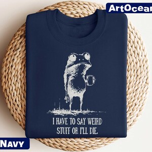 A navy blue t-shirt with a white illustration of a frog and the text "I HAVE TO SAY WEIRD STUFF OR ILL die" printed on it.