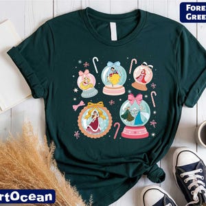 A green t-shirt with a colorful graphic design featuring snowglows and various characters, including a princess, a fairy, and a snowman. The t-shirt is laid out on a wooden surface, along with some books, a cup, and a pair of sneakers.