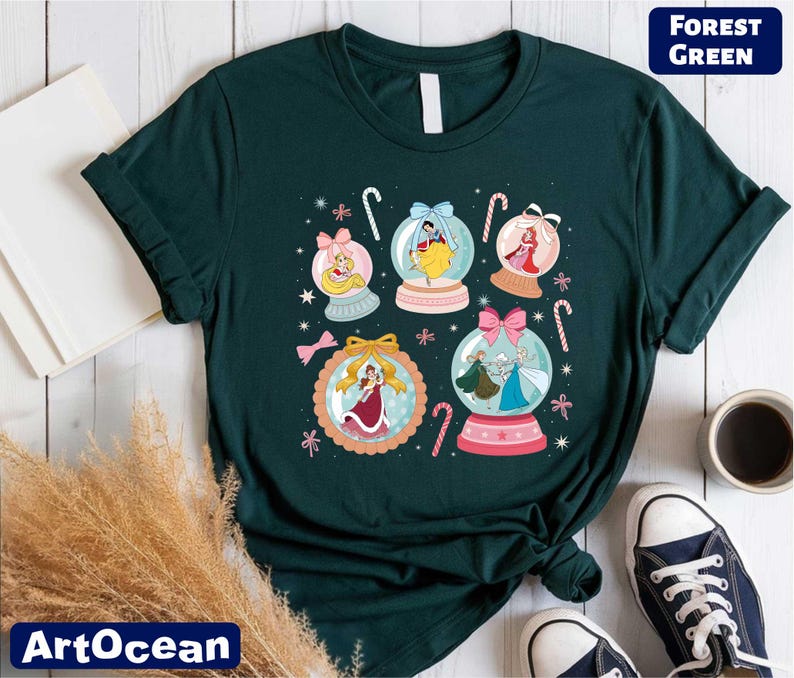A green t-shirt with a colorful graphic design featuring snowglows and various characters, including a princess, a fairy, and a snowman. The t-shirt is laid out on a wooden surface, along with some books, a cup, and a pair of sneakers.