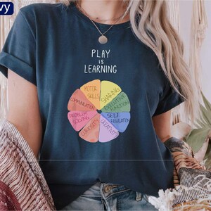 A person wearing a navy blue t-shirt with a colorful wheel graphic and the text "PLAY IS LEARNING" printed on it.