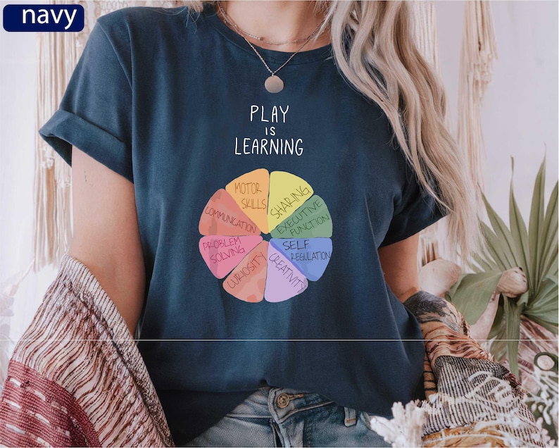 A person wearing a navy blue t-shirt with a colorful wheel graphic and the text "PLAY IS LEARNING" printed on it.