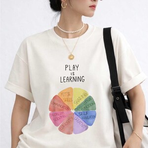 A woman wearing a white t-shirt with a colorful wheel graphic and the text "PLAY IS LEARNING" printed on it, standing against a white background.