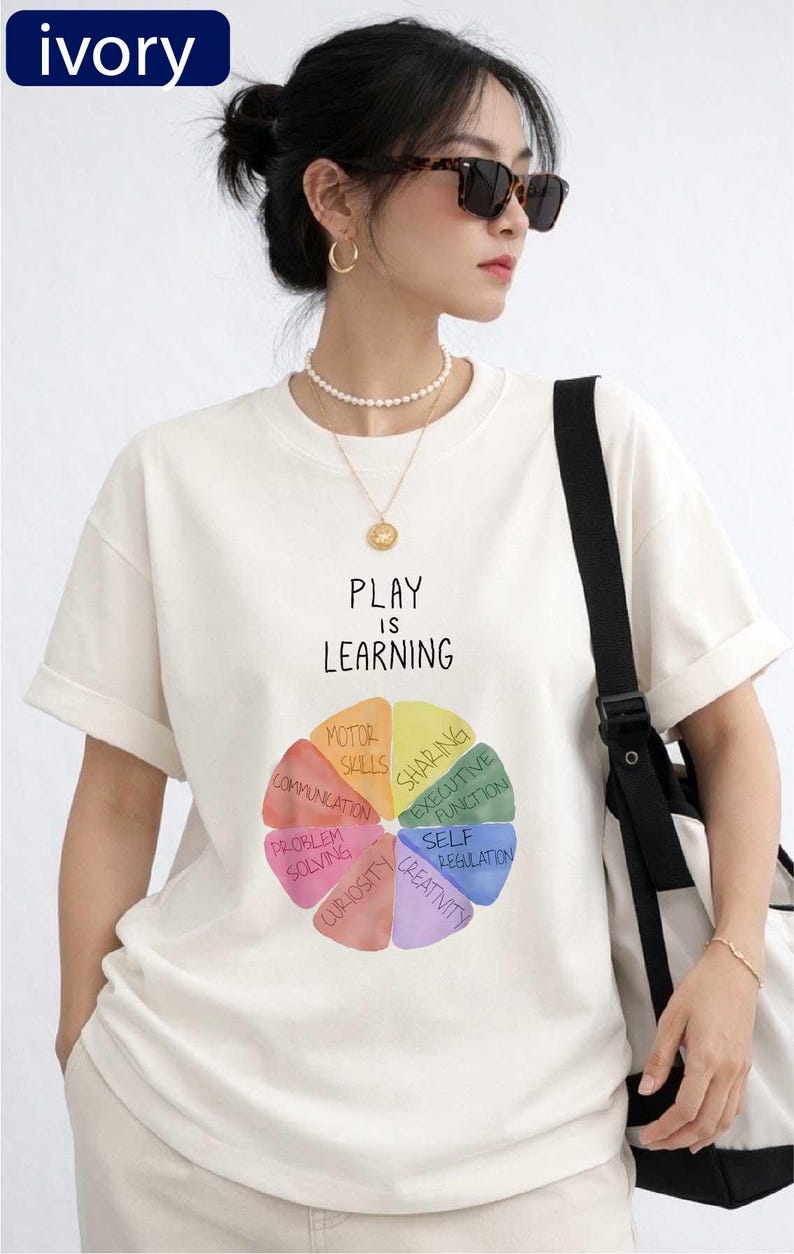 A woman wearing a white t-shirt with a colorful wheel graphic and the text "PLAY IS LEARNING" printed on it, standing against a white background.