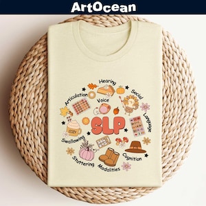 May include: A beige t-shirt with a graphic of a Thanksgiving turkey and fall-themed elements surrounding the word "SLP". The graphic includes words like "Hearing", "Voice", "Articulation", "Swallowing", "Stuttering", "Modalities", "Cognition", "Social", and "Language".
