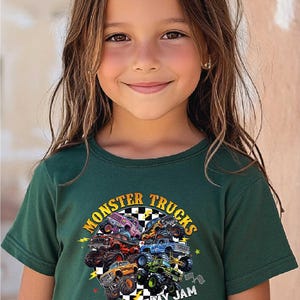 A young girl with long brown hair is smiling and wearing a green t-shirt with a graphic of monster trucks and the text "Monsters Trucks Are My Jam".