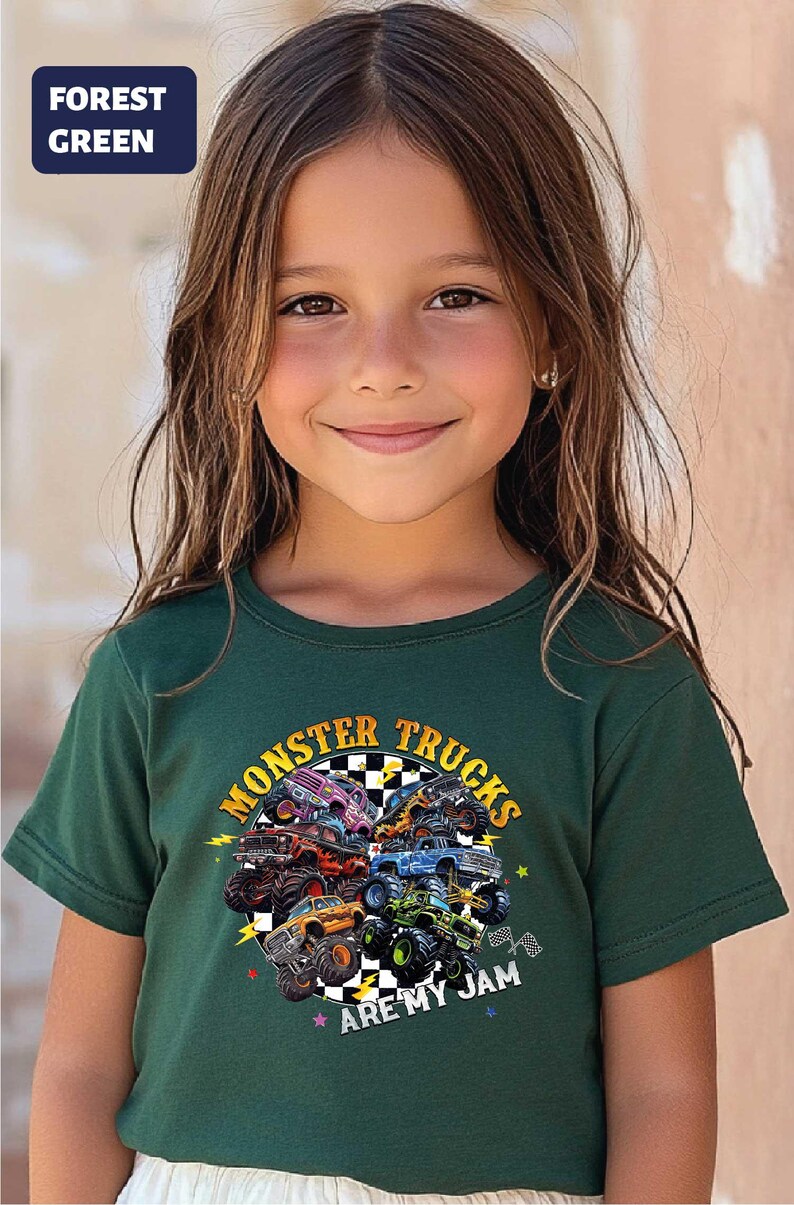 A young girl with long brown hair is smiling and wearing a green t-shirt with a graphic of monster trucks and the text "Monsters Trucks Are My Jam".