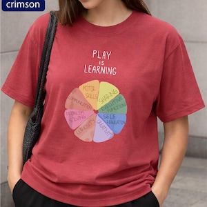 A woman wearing a red t-shirt with a colorful wheel graphic and text that says "PLAY IS LEARNING".