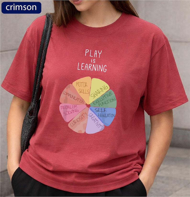 A woman wearing a red t-shirt with a colorful wheel graphic and text that says "PLAY IS LEARNING".