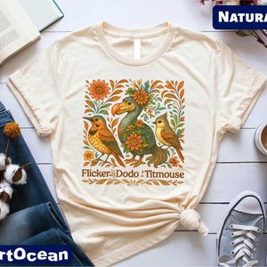 May include: A beige t-shirt with a vintage-style illustration of a dodo bird, a flicker bird, and a titmouse. The birds are surrounded by flowers and leaves. The text "Flicker Dodo Titmouse" is printed below the illustration.