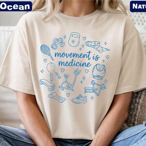 A person wearing a white t-shirt with a graphic design featuring various sports and fitness-related icons, along with the text "movement is medicine".
