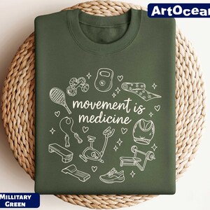 A green t-shirt with a white graphic design featuring various fitness-related objects such as a tennis racket, dumbbells, and a yoga mat, along with the text "motion is medicine".