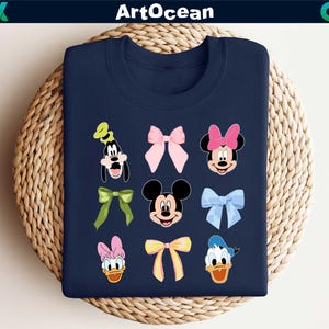 May include: Navy blue sweatshirt featuring cartoon character faces and bows. Characters include Goofy, Minnie Mouse, Mickey Mouse, Donald Duck, and Daisy Duck. The word "ArtOcean" is at the top of the sweatshirt.