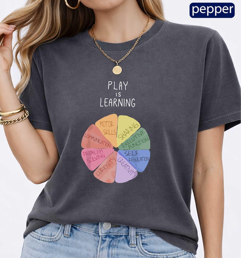 A woman wearing a gray t-shirt with a colorful wheel graphic and the text "PLAY IS LEARNING" printed on it.