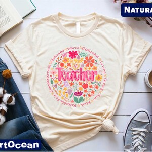 May include: A cream colored t-shirt with a floral design and the word "Teacher" in the center. The design includes the words "Warm, Dedicated, Loving, Caring, Fun, Kind, Patient, Believing, Compassionate, Motivator" around the floral design. The shirt is labeled "NATURAL".