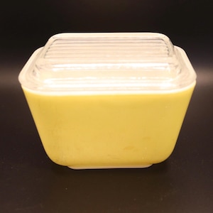 Puede incluir: Una fuente de horno cuadrada de vidrio amarillo vintage con una tapa de vidrio transparente y acanalado. El plato tiene una forma ligeramente cónica y un borde blanco. La tapa encaja perfectamente, creando un artículo de cocina completo y funcional.