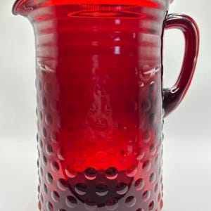 May include: A ruby red glass pitcher with a handle and a spout. The pitcher has a textured pattern of small, raised circles across its surface. The rim of the pitcher is smooth and the handle is curved. A classic piece for serving beverages.