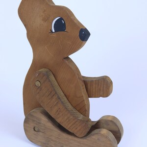 May include: A wooden toy bear with painted eyes and a black nose. The bear is articulated at the arms and legs, allowing it to move.