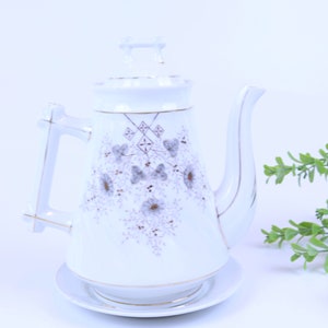 May include: White ceramic teapot with a matching lid and saucer. The teapot features a floral design in shades of gray and brown, with gold trim accents. The handle and spout are elegantly shaped.