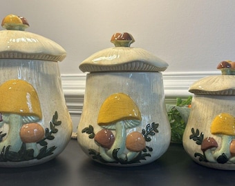 May include: A set of three ceramic canisters with lids shaped like mushroom caps. Each off-white canister features a raised, colorful mushroom design in yellow, brown, and orange with green foliage accents. The lids have a small mushroom finial.