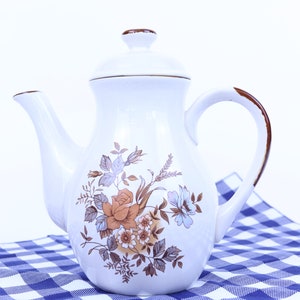 May include: A white ceramic teapot with a floral design in shades of brown, orange, and blue. The teapot has a brown rim and a brown handle. It is sitting on a blue and white checkered tablecloth.