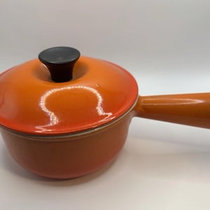 May include: An orange enameled cast iron saucepan with a lid and a single handle. The lid has a black knob. The cookware is designed for stovetop use. The pan is a vibrant orange colour.