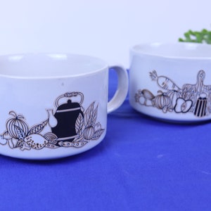May include: Two white ceramic soup bowls with black and brown illustrations of vegetables and a teapot. The bowls have a rounded shape and a single handle.